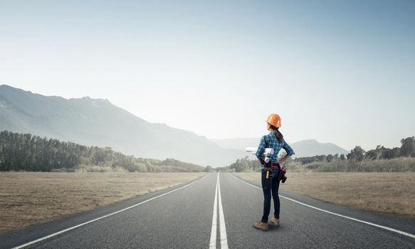 Young woman in safety helmet standing on asphalt road with blueprints. Back view of female architect in workwear checking new construction site at countryside. Road infrastructure reconstruction.