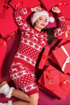 Funny young woman in a New Year's dress lying on a mountain of gifts. Christmas sale. Emotional brunette in Santa hat