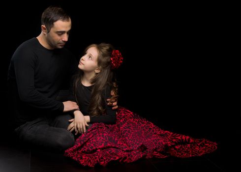 Portrait of Loving Father Hugging His Beautiful Daughter, Handsome Young Man Sitting on the Floor with his Adorable Girl, Happy Family Sitting Against Black Studio Background