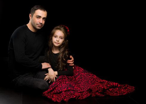 Portrait of Loving Father Hugging His Beautiful Daughter, Handsome Young Man Sitting on the Floor with his Adorable Girl, Happy Family Sitting Against Black Studio Background