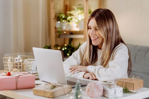 Happy and smiling young woman wrapping and preparing Christmas gifts, typing at laptop and having online video chat. Online shopping at Christmas holidays. Home interior with lights and plants