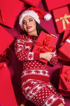 Funny young woman in a New Year's dress lying on a mountain of gifts. Christmas sale. Emotional brunette in Santa hat