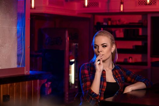 Young pretty woman in a shirt in a cage smoke an electronic cigarette at the vape bar. Vape shop.