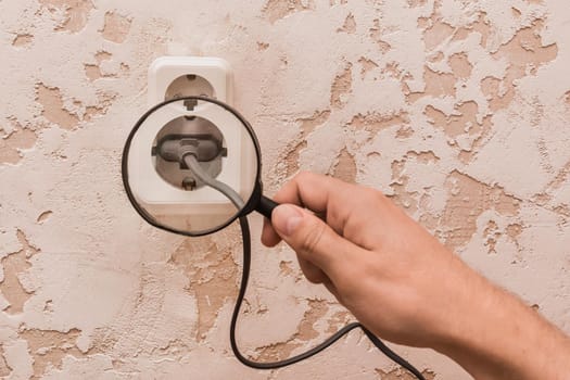 The hand of a working man with a magnifying glass examines a cord connected to a double socket. The concept of keeping household appliances safe.