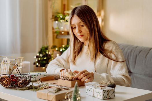 Woman wrapping Christmas eco natural gift boxes at home. Preparing presents on white table with decor elements, lights and items Christmas or New year DIY packing Concept.