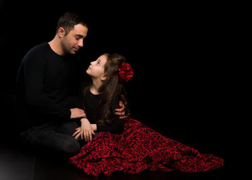 Portrait of Loving Father Hugging His Beautiful Daughter, Handsome Young Man Sitting on the Floor with his Adorable Girl, Happy Family Sitting Against Black Studio Background