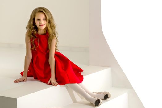 Pretty Girl Wearing Nice Red Dress Sitting on White Stairs, Portrait of Preteen Girl with Joyful Face Expression, Charming Fashionable Child Dressed Stylish Dress Posing in Studio Interior