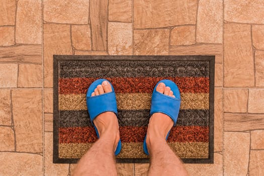 Male feet in blue house slippers stand on a foot mat on a brown tiled floor texture background, top view.