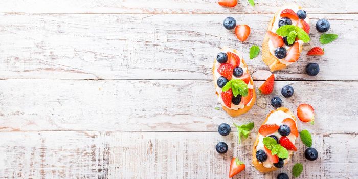 Background with crostini with grilled baguette, cream cheese, blueberry, strawberry, honey and mint on old white wooden background. Delicious appetizer or dessert. Copy space.