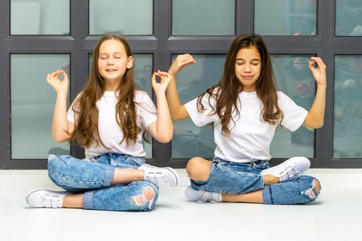 Two cute little girls meditate near the window, they are sitting in the lotus position. The concept of tenderness and beauty.