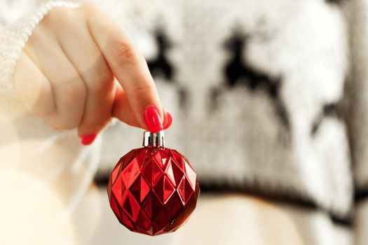 Hand of a woman in festive sweater holding Christmas bauble with blurred lights close up