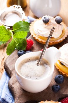 Cup of coffee and delicious homemade tartlets served with lemon, lime curd cream, berries and meringue on old cutting board.