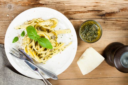 Cooked homemade tagliatelle pasta with green pesto sauce, grated pecorino cheese and basil on white plate on old wooden background. Top view.