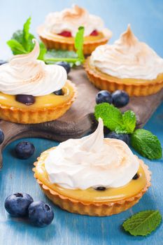 Handmade little tarts, cakes, tartlets with lime curd, meringue en berries on old rustik board on blue wooden background. Selective focus.