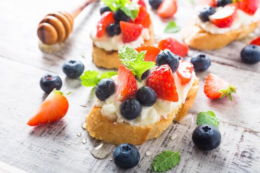 Small canape, crostini with grilled baguette with cream cheese, blueberry, strawberry, honey and mint on old white wooden background. Delicious appetizer or dessert.