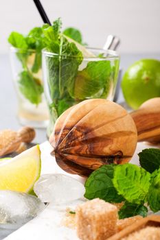 Wooden juicer and ingredients for making mojitos. Ice, fresh mint leaves sugar cubs, lime and glass on white cutting board
