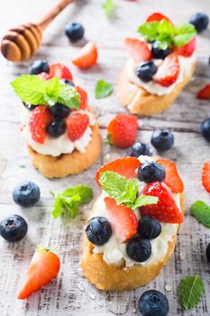 Small canape, crostini with grilled baguette with cream cheese, blueberry, strawberry, honey and mint on old white wooden background. Delicious appetizer or dessert.