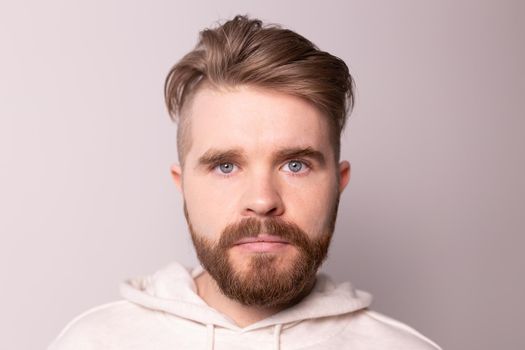 Portrait of young male with beard and mustache and trendy hairdo. Wears casual hoodie, has serious expression in studio against white