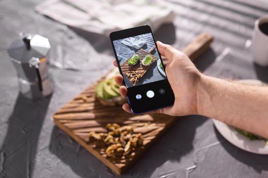 Hands take pictures on smartphone of two beautiful healthy sour cream and avocado sandwiches lying on board on the table. Social media and food