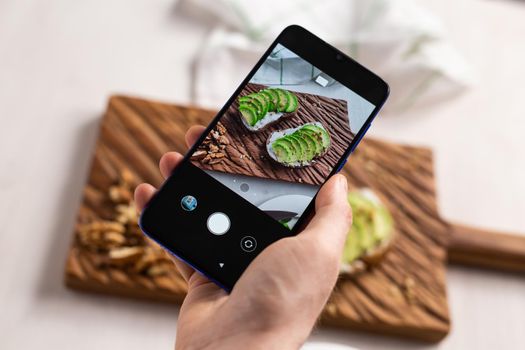 Hands take pictures on smartphone of two beautiful healthy sour cream and avocado sandwiches lying on board on the table. Social media and food