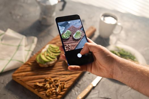 Hands take pictures on smartphone of two beautiful healthy sour cream and avocado sandwiches lying on board on the table. Social media and food