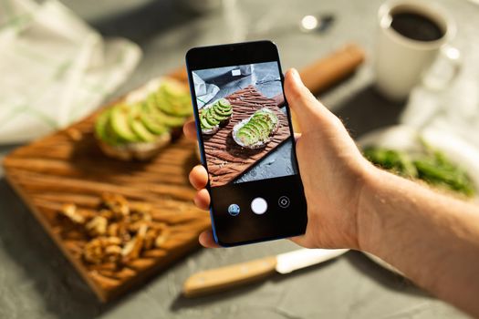 Hands take pictures on smartphone of two beautiful healthy sour cream and avocado sandwiches lying on board on the table. Social media and food