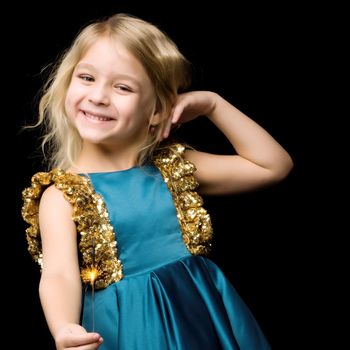 Beautiful happy girl holds a burning sparkler, a charming girl in a beautiful blue dress plays fiery sparklers in the dark, portrait of a cute stylish blonde isolated on black background.Close-up.