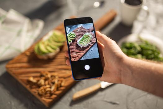 Hands take pictures on smartphone of two beautiful healthy sour cream and avocado sandwiches lying on board on the table. Social media and food