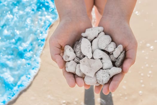 The hands of a young girl close-up hold a pile of white stones against the backdrop of the sea coast.