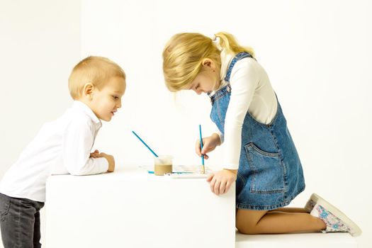 Cute Blonde Girl Sitting on Her Knees and Painting with Paintbrush, Her Brother Looking at Her with Interst, Adorable Brother and Sister in Fashionable Clothes, Portrait of Cheerful Stylish Children