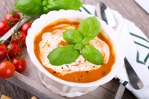 Fresh tomato soup with basil and cream in a white bowl on wooden background. Selective focus.