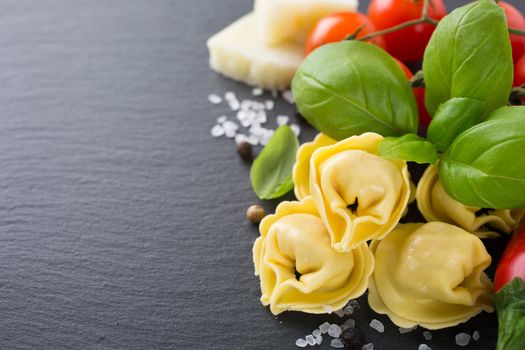 Homemade raw Italian tortellini filled with ham and cheese with raw tomatoes, basil leaves and cheese on black background. Healthy food concept with copy space concept. Selective focus.