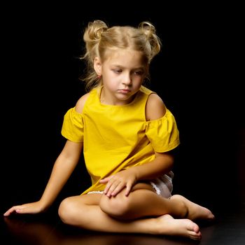 Beautiful little girl is sitting on the floor in the studio. The concept of a happy childhood, beauty and fashion. Isolated over black background.