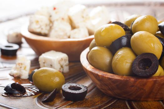 Fresh green and black olives in olive wood bowl and feta cheese on rustic wooden background. Selective focus.