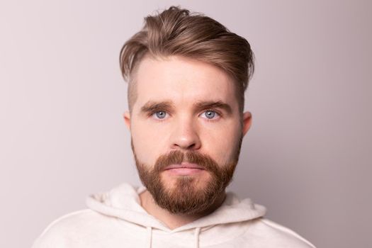 Portrait of young male with beard and mustache and trendy hairdo. Wears casual hoodie, has serious expression in studio against white
