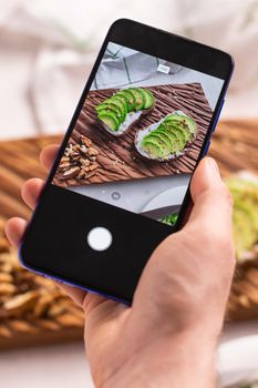 Hands take pictures on smartphone of two beautiful healthy sour cream and avocado sandwiches lying on board on the table. Social media and food