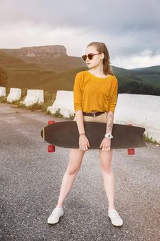 Beautiful and fashionable young woman in sunglasses and with a tattoo poses with a skateboard or longboard against the sunset sky.