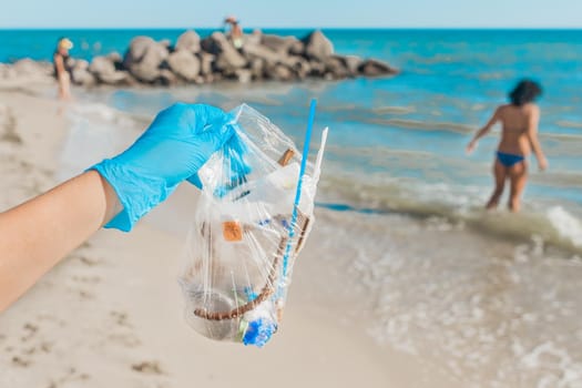 A man's hand in protective latex gloves holds a bag of waste against the sea. Cleaning up the garbage behind people.