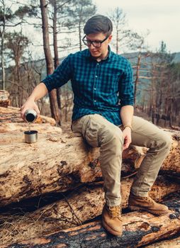 Traveler young man sitting on fallen tree trunk and pouring tea from thermos to cup in the forest. Man has a picnic outdoor