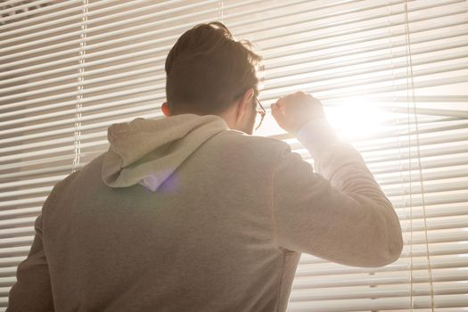 Rear view of young stylish man with beard and coffee peeking through hole in window blinds and looking out into street. Enjoying the morning sun and positivity