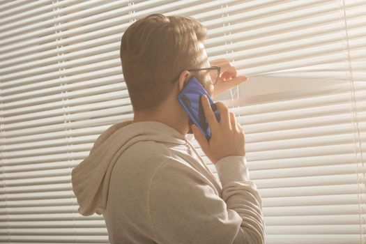 Pensive guy is talking on his smartphone and looking at window at home. Male thinking dreaming and looking out window.