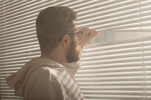 Rear view of young man with beard peeks through hole in the window blinds and looks out into the street. Surveillance and curiosity