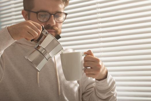 Close-up portrait of young stylish hipster male pouring coffee at office on summer day. Invigorating morning and positive mood