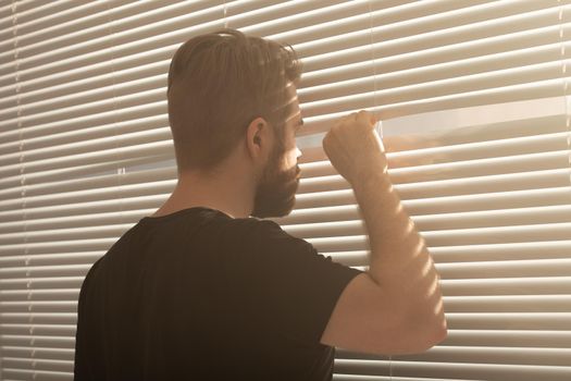 Rear view of young man with beard peeks through hole in the window blinds and looks out into the street. Surveillance and curiosity