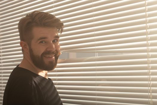 Rear view of young man with beard peeks through hole in the window blinds and looks out into the street. Surveillance and curiosity