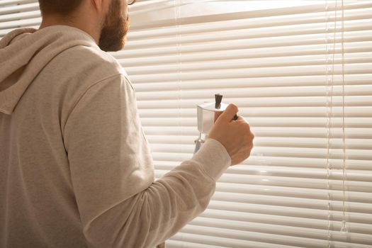 Rear view of young stylish man with beard and coffee peeking through hole in window blinds and looking out into street. Enjoying the morning sun and positivity