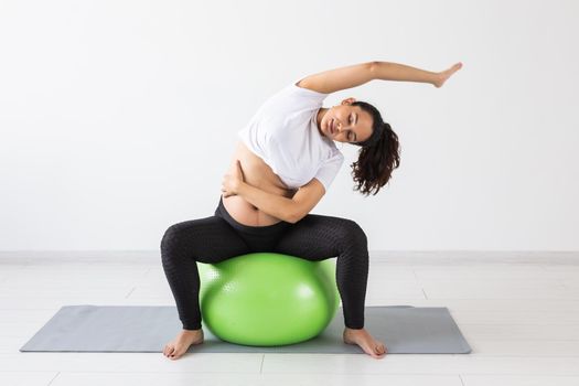 A young pregnant woman doing relaxation exercise using a fitness ball while sitting on a mat and holding her tummy