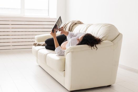 Latin pregnant woman using tablet computer sitting on sofa at home. Pregnancy and information for parenthood