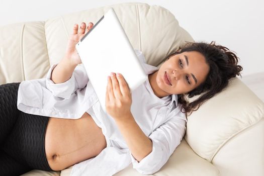 Latin pregnant woman using tablet computer sitting on sofa at home. Pregnancy and information for parenthood