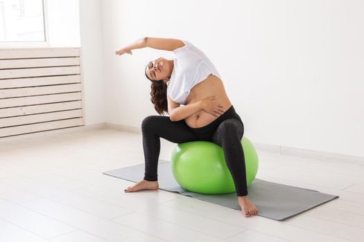 A young pregnant woman doing relaxation exercise using a fitness ball while sitting on a mat and holding her tummy